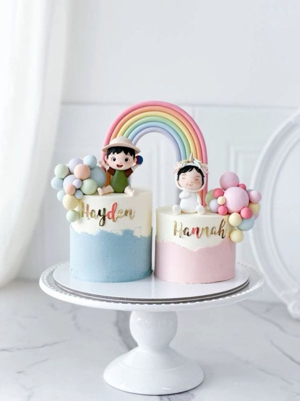 Pastel twin birthday cake with rainbow arch, fondant baby toppers, and name personalization for boy and girl twins